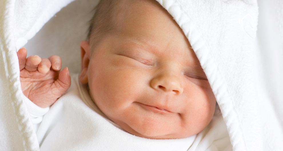 A close-up of a newborn baby's face, partially covered by a white cloth, with the baby's hand raised.