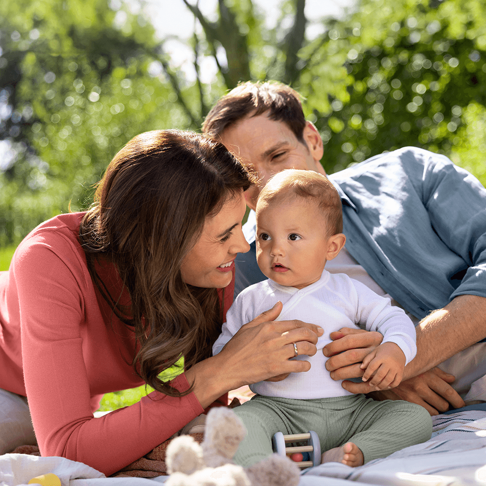 A man and a woman lie on a blanket outdoors, engaging with a baby.