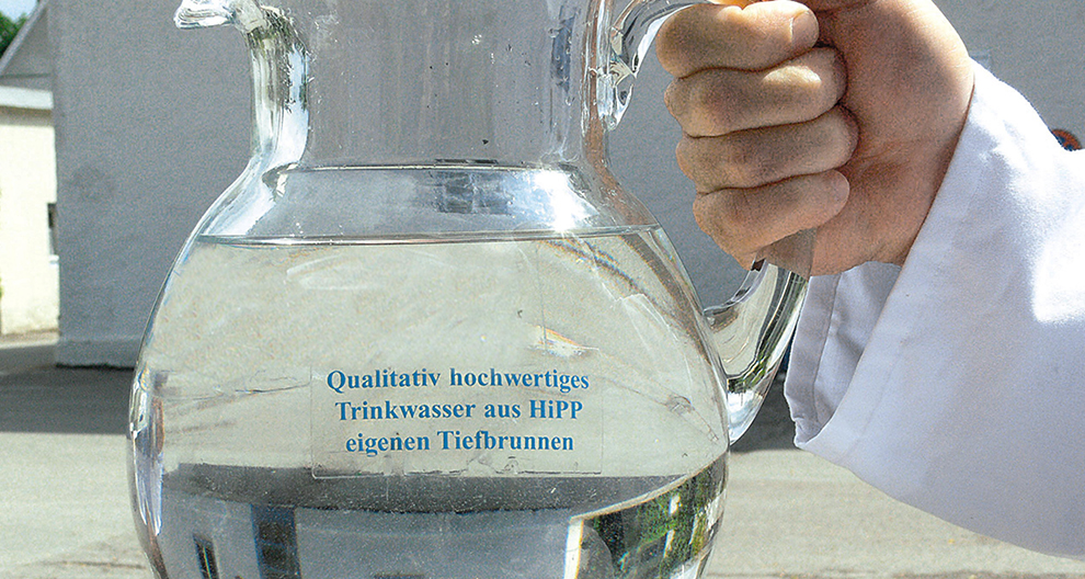 A hand in a white sleeve holds a clear glass jug filled with water, with a label in German on the jug.