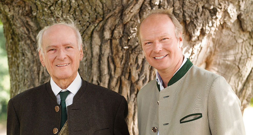 Two men, one older and one younger, dressed in formal attire, stand side by side against the textured bark of a tree.
