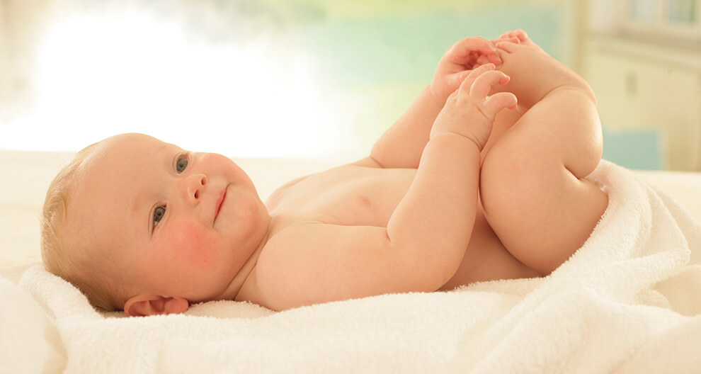 A baby lies on a white blanket, holding its foot.