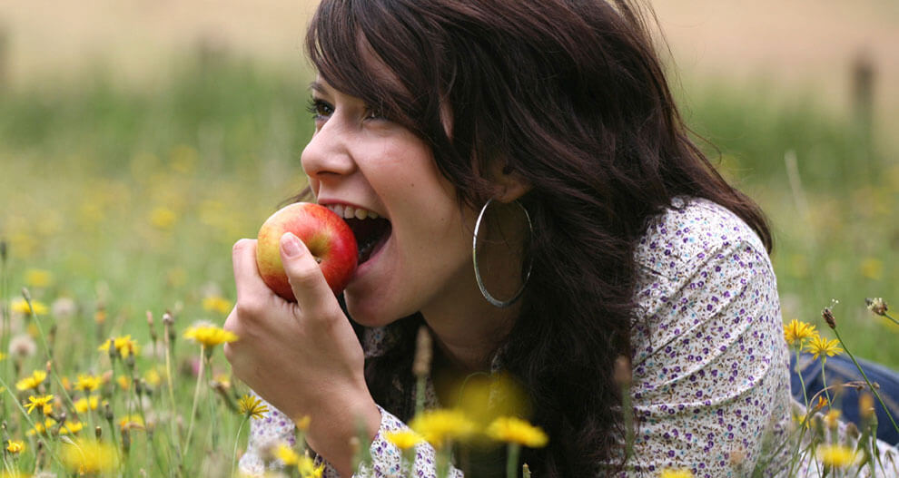 A woman with long dark hair is lying in a field of yellow flowers, biting into a red apple.