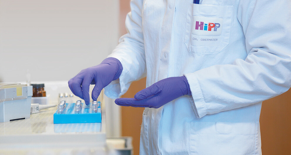 A person in a white lab coat with the logo "HiPP" and the name "CH. OBERMAIER" is wearing purple gloves and handling a blue tray with multiple small containers.