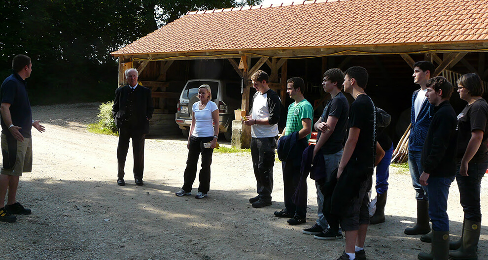 A group of people stands in a line outside a barn, engaged in a conversation.