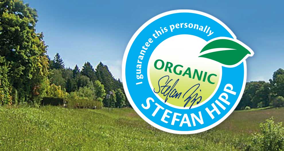Pokrajina z drevesi in travo je prekrita s krožno grafiko z napisom "Za to jamčim osebno" in "Stefan Hipp" s simbolom lista in besedo "Organic".