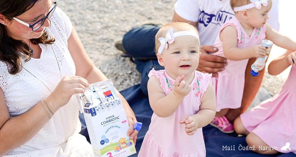 Ženska v beli obleki drži škatlo "Junior Combiotic", medtem ko trije malčki v rožnatih oblekah sedijo na modri odeji, eden od njih pa drži modro stekleničko.