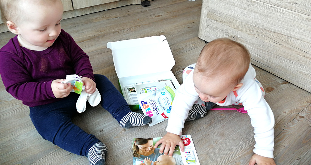 Na lesenih tleh sedita dva malčka, eden drži pisan paket, drugi pa seže po reviji z žensko in dojenčkom na naslovnici.