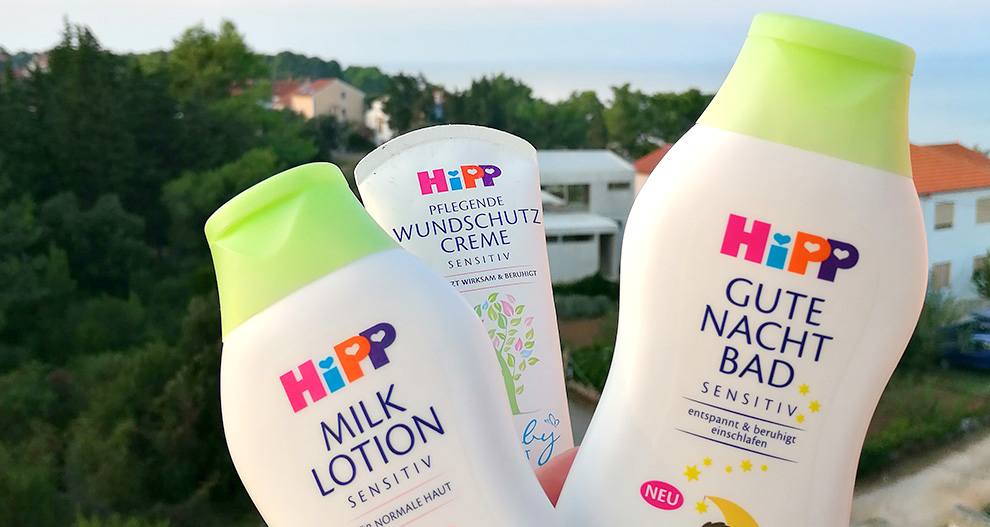 Tri bele stekleničke z zelenimi pokrovčki z napisi "MILK LOTION", "WUNDSCHUTZ CREME" in "GUTE NACHT BAD" stojijo na zamegljenem ozadju dreves, hiš in vodnega telesa.