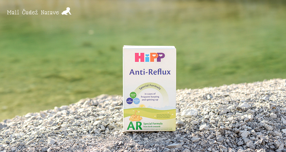 Bela škatla z napisom "HiPP Anti-Reflux" z zelenim in modrim vzorcem z risanim medvedom je postavljena na posteljo iz sivih kamnov z zamegljenim zelenim ozadjem.