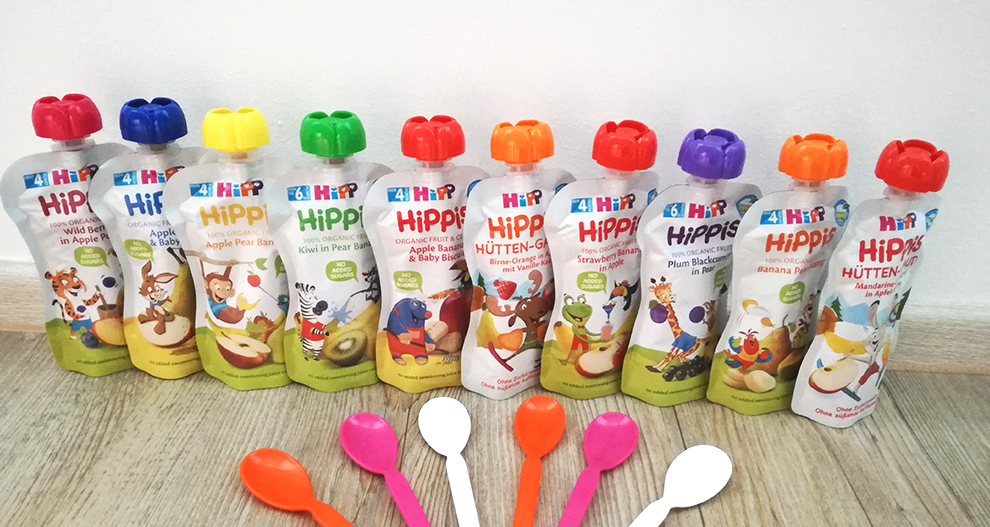 Niz osmih vrečk za ekološko otroško hrano "HiPP" z različnimi kombinacijami sadja, ki jih spremljajo tri plastične žlice v roza, oranžni in beli barvi.