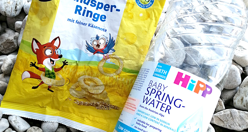Rumena vrečka za malico z ilustracijo lisice in ptice ter prozorna plastenka z napisom "Hipp Baby Spring Water" sta postavljeni na podlago iz kamenčkov.