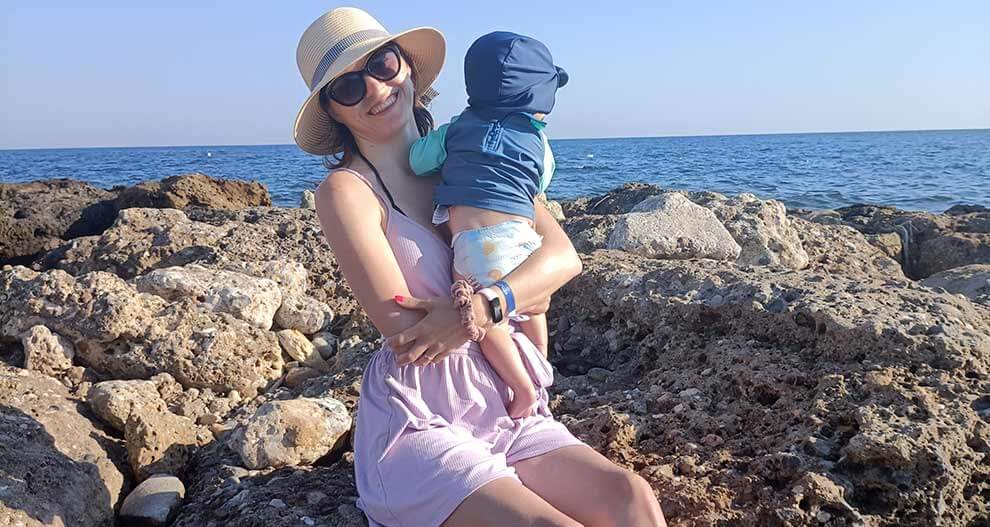 Ženska v rožnati obleki in slamnatem klobuku sedi na skalnatem terenu in drži otroka v modri majici in beli plenički.