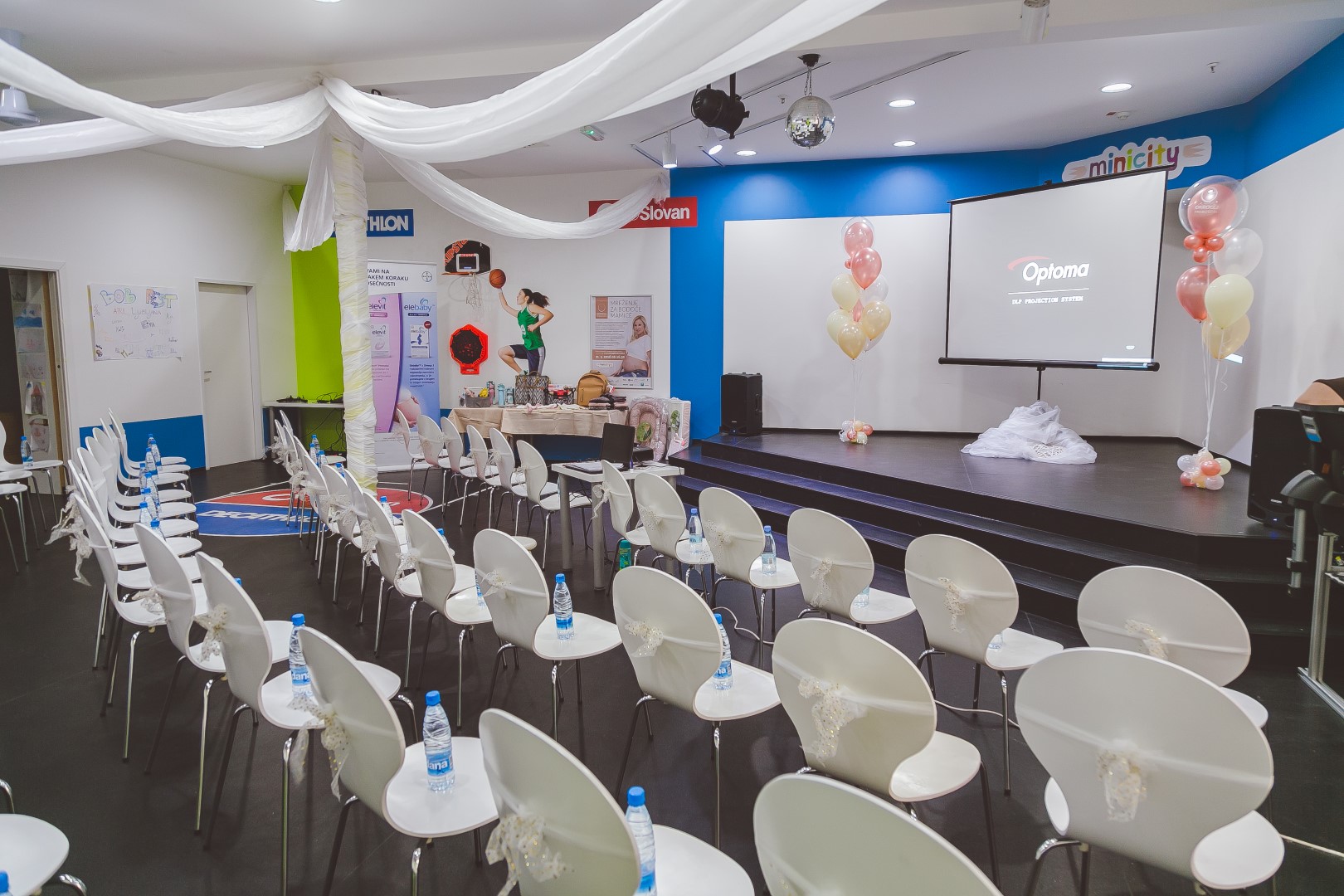 V konferenčni sobi so postavljene vrste belih stolov, projekcijsko platno z logotipom Optoma ter oder z baloni in belo tkanino.