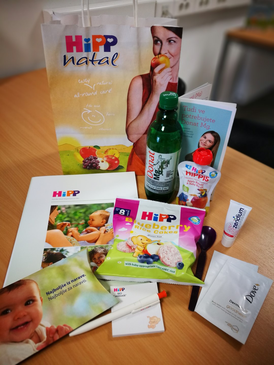 Na mizi so razstavljeni različni izdelki in materiali, povezani z dojenčki, vključno z nakupovalno vrečko, hrano, stekleničko in informativnimi zloženkami.