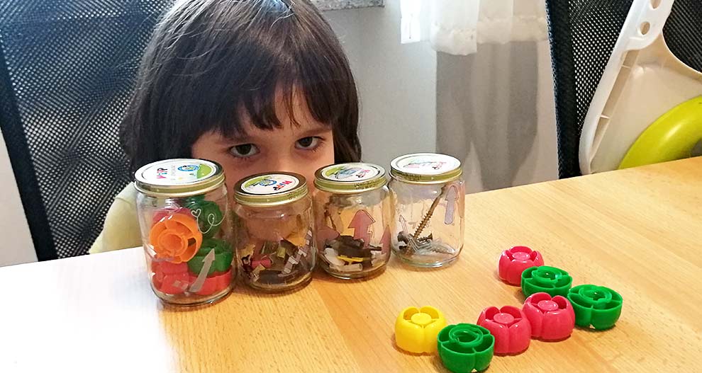 Otrok sedi za mizo s štirimi steklenimi kozarci z različnimi predmeti, pred njimi pa so raztresene plastične igrače v obliki cvetov.