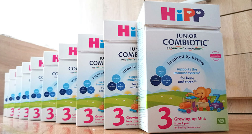 Na lesenih tleh je poravnanih več belih škatel s pisanimi grafikami in besedilom, vsaka z napisom "Hipp Junior Combiotic" in "Rastoče mleko od 1. leta starosti za zdrav razvoj".
