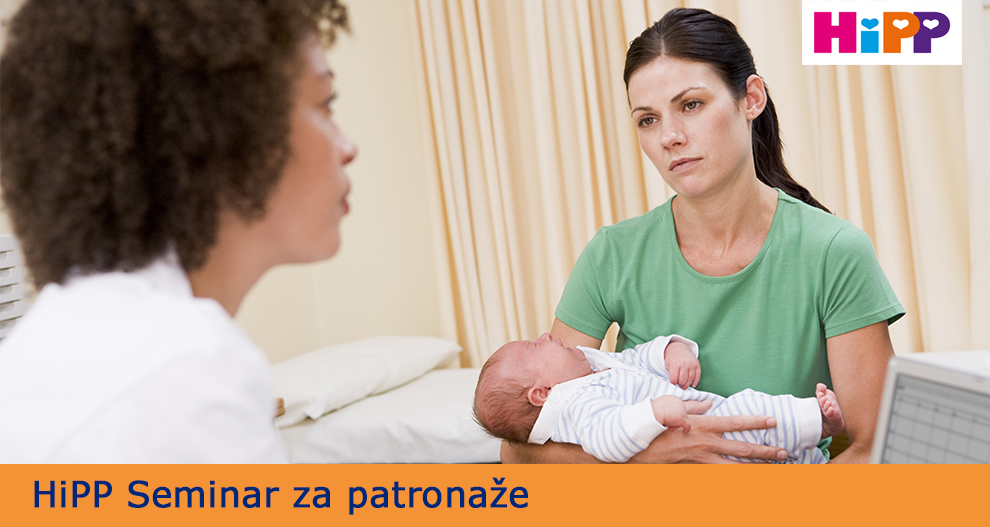 Ženska v zeleni srajci drži dojenčka, sedi v sobi z zaveso, medtem ko ji govori druga oseba, katere obraz ni povsem viden.